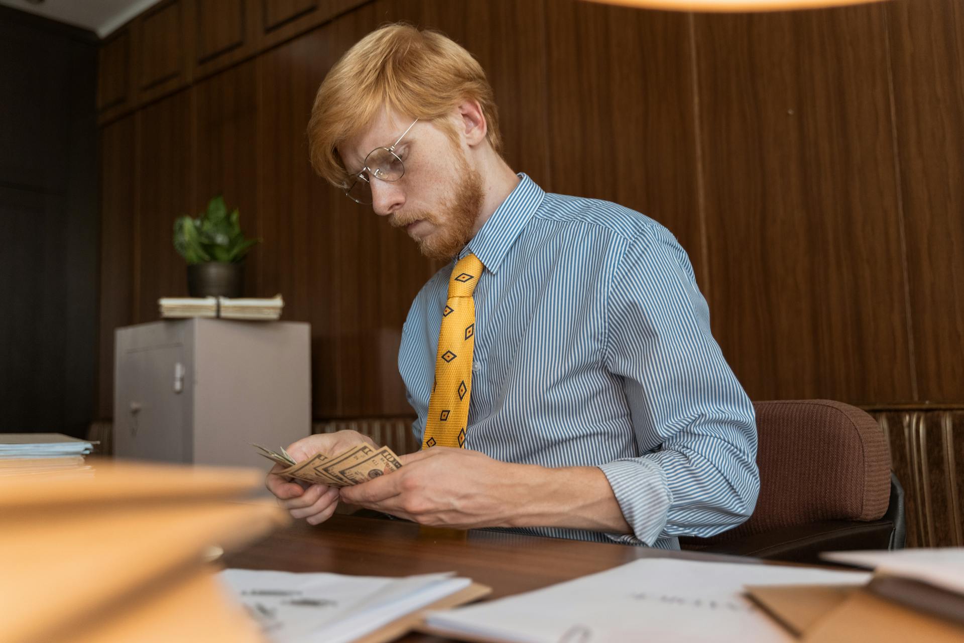 Serious businessman counting cash at his desk in an office environment with wood paneling.