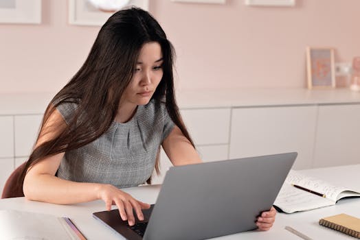 Asian woman in a modern office setting working on a laptop, symbolizing entrepreneurship and professionalism.