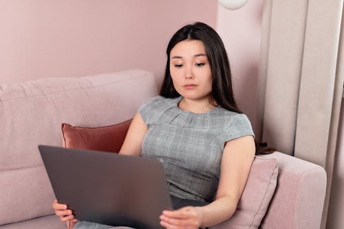 Free Woman in a Gray Dress Looking at Her Laptop while Sitting on a Sofa Stock Photo