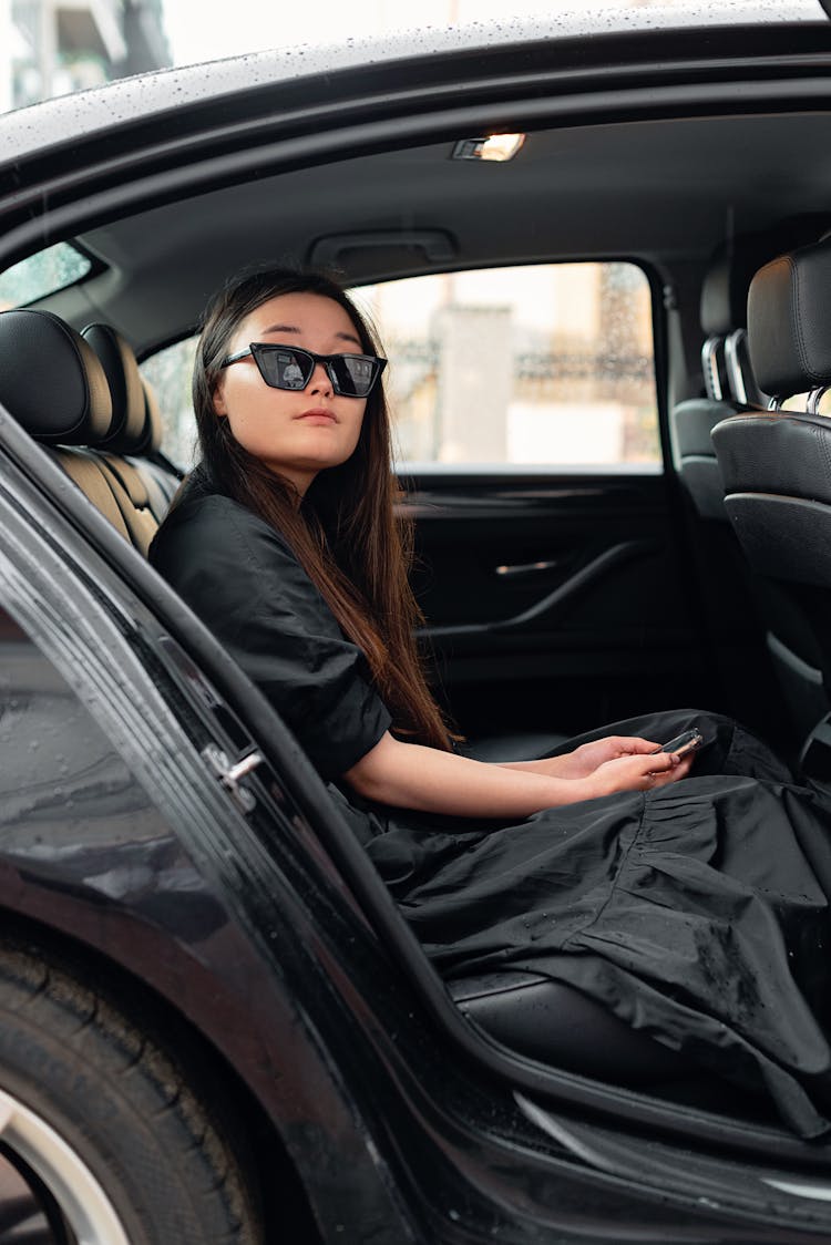 A Woman In Black Dress Wearing Black Framed Sunglasses Sitting In A Car