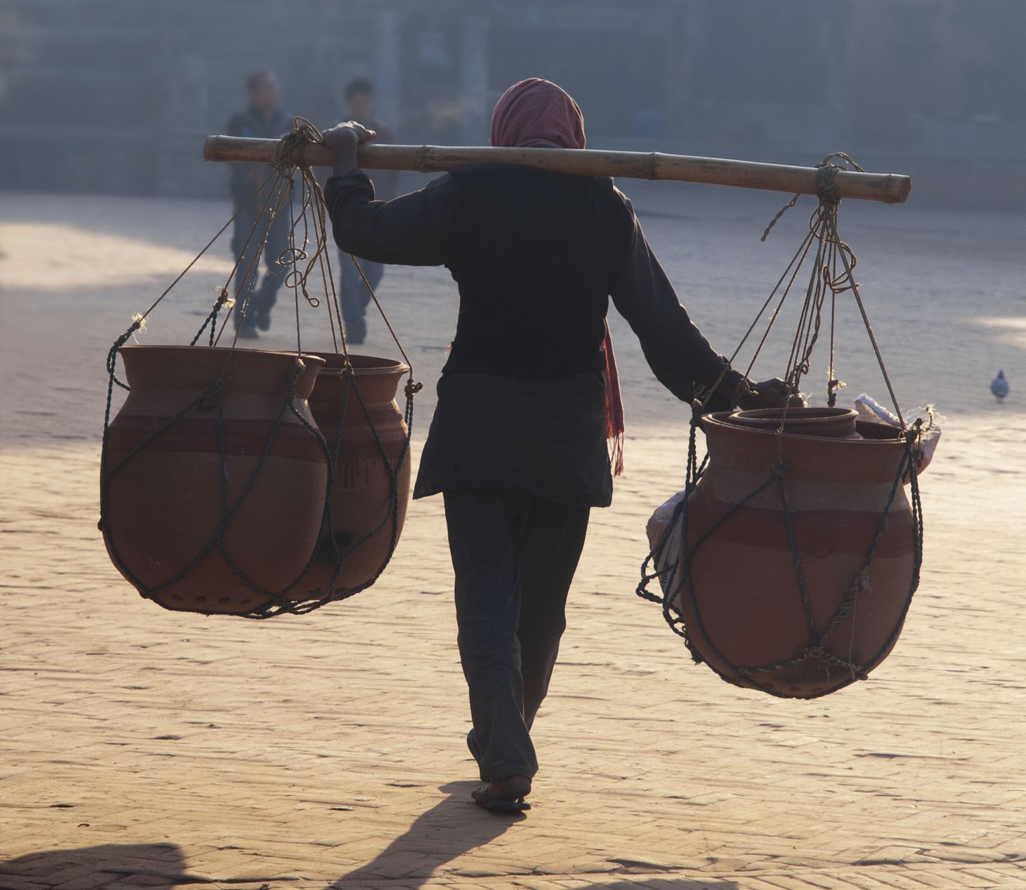 Back view of a Nepalese merchant carrying clay pots with a shoulder pole in the morning light.