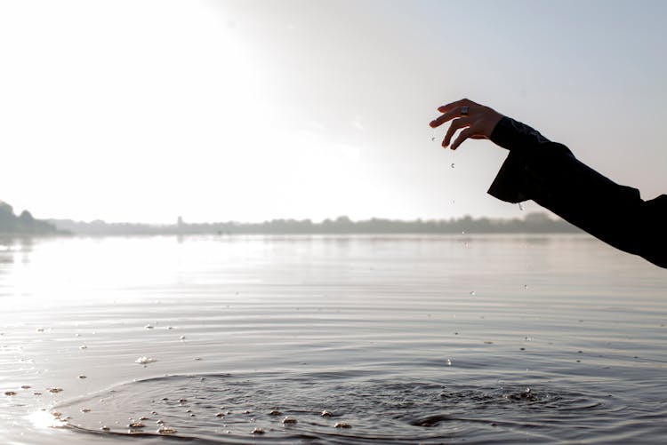 Person's Hand With Water Droplets Forming Ripples On Water Surface