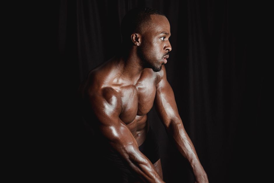 A fit and muscular black man striking a powerful pose shirtless in a dark studio setting.