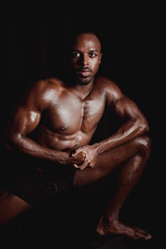 Fit and muscular man posing shirtless against a dark background, showcasing defined muscles.