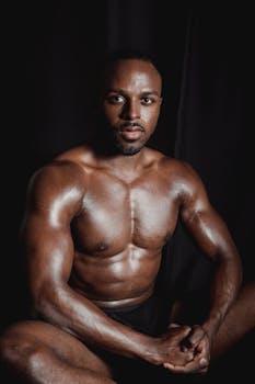 A powerful portrait of a muscular male sitting shirtless against a dark background, showcasing strength.