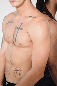 Two shirtless tattooed men standing back to back in a studio setting with a white background.