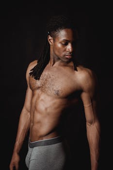A muscular, shirtless man with dreadlocks poses against a black background in a studio setting.