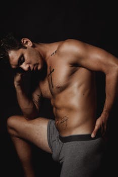 Muscular man posing in a dramatic studio setting, showcasing tattoos and physique.