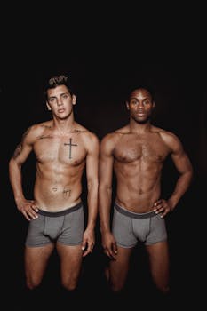 Two shirtless men posing confidently in a studio setting, showcasing tattoos and muscle definition.