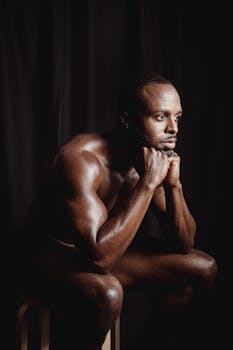 A shirtless man with a muscular build poses pensively against a dark background.