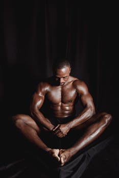 Shirtless bodybuilder sitting against a dark backdrop, highlighting muscular physique.