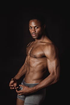A muscular black man poses shirtless against a dark background, showcasing fitness and strength.