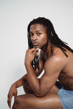 Studio portrait of a fit, shirtless man with dreadlocks, sitting against a white background.
