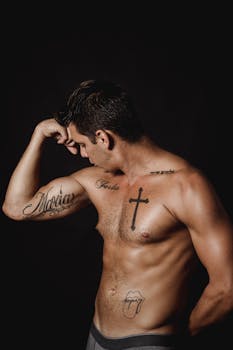 Shirtless man with tattoos flexes bicep against a dark background, showcasing muscular physique.