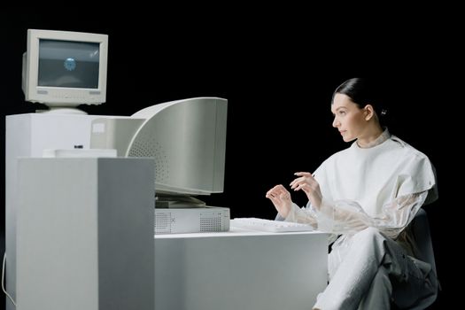 A woman in a futuristic outfit interacts with a vintage computer in a minimalist environment.
