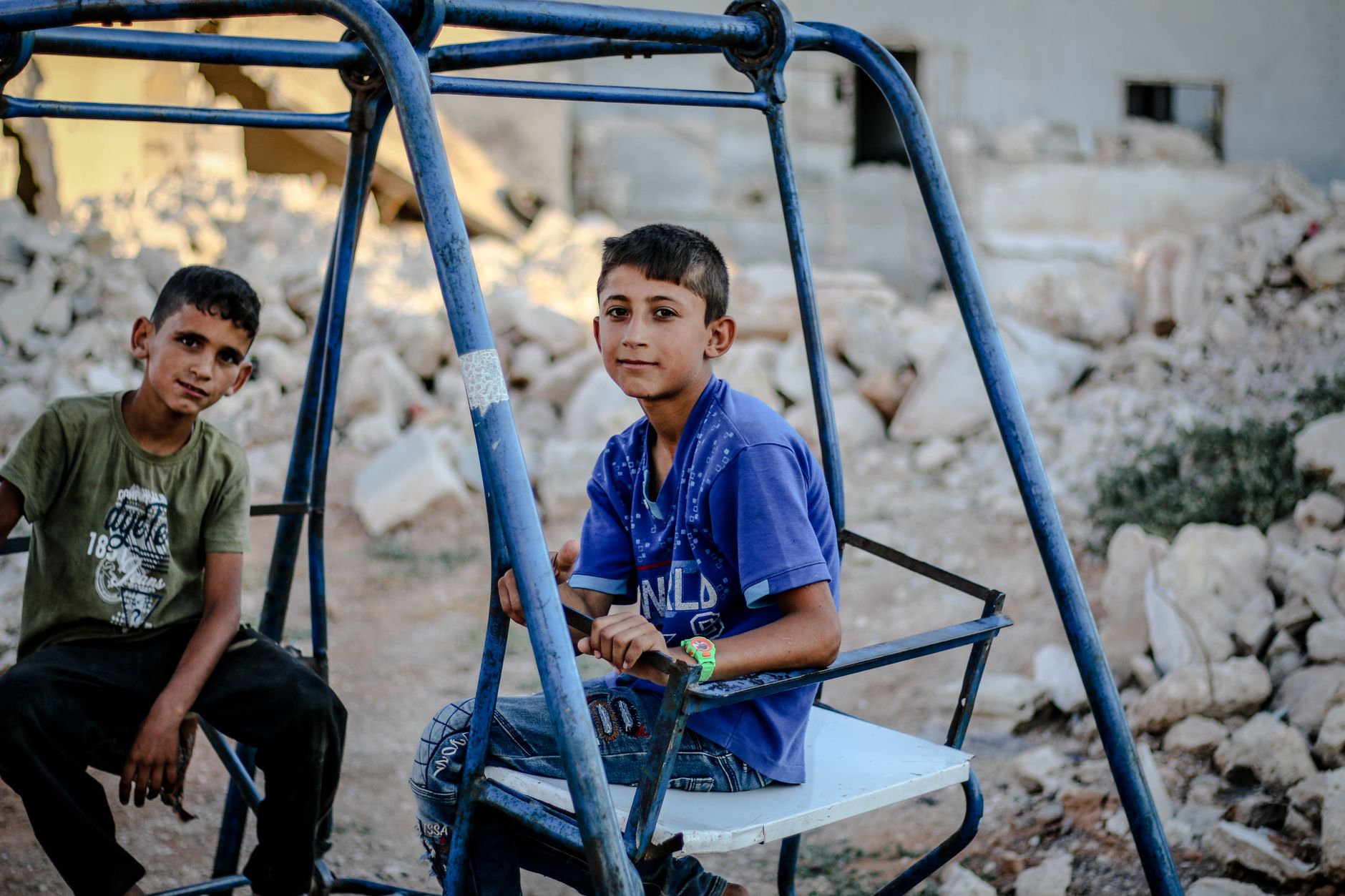 Twee jongens zittend op een schommel in de open lucht te midden van puin in Idlib, Syrië, wat levensveerkracht uitbeeldt.