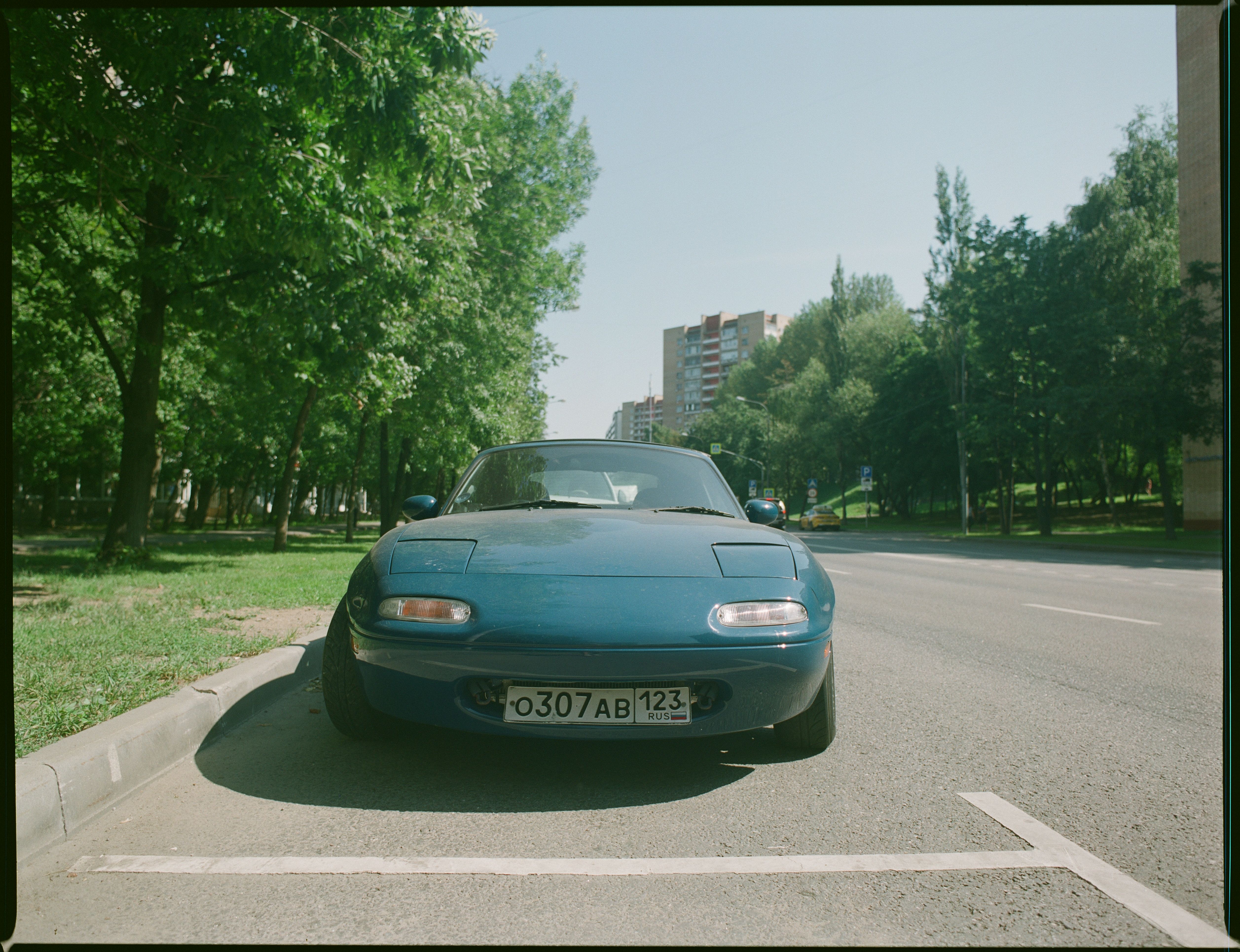 Classic blue car parked on a sunny urban street with lush trees.