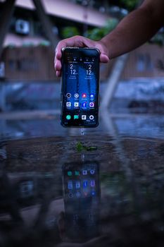 Hand holding a smartphone above a reflective wet surface with apps visible, outdoors.