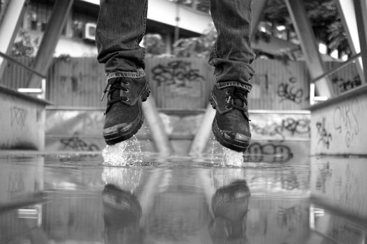 Monochrome image capturing a pair of shoes splashing water midair, reflecting in a puddle.