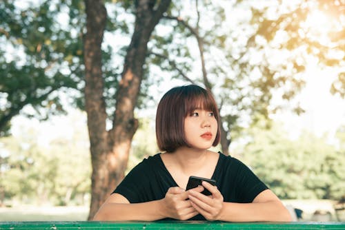Free Woman Holding Smartphone Wearing Black Shirt Standing Under Tree Stock Photo