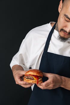 A chef in an apron holds a juicy burger with ketchup in a studio setting.