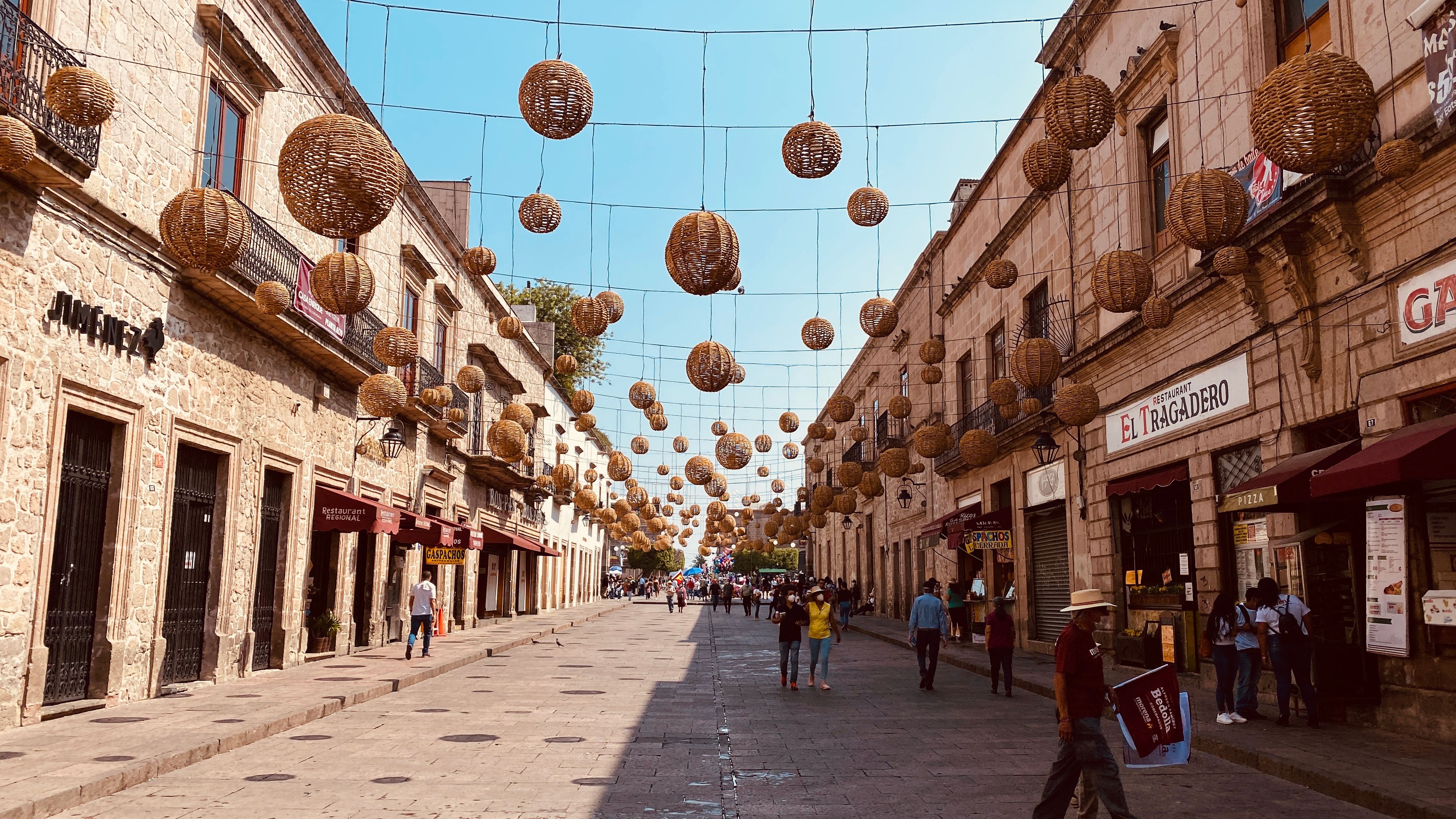 Street view of Morelia, Mexico with hanging woven lanterns and pedestrians on a sunny day.
