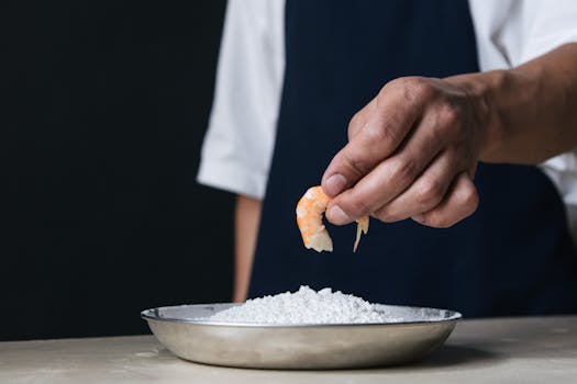 Close-up of a chef's hand holding shrimp above a stainless bowl with flour.