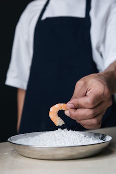 Close-up of a chef's hand holding shrimp over a bowl of flour, symbolizing food preparation.