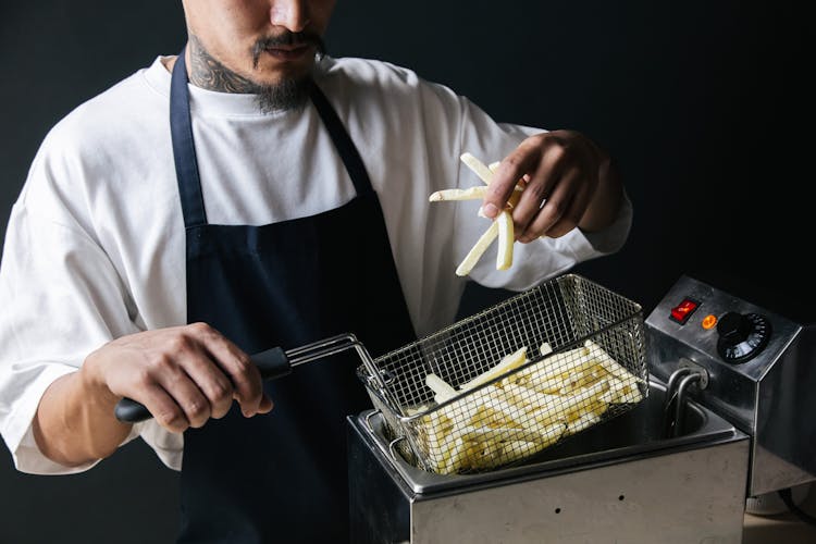 A Man In White Shirt With Black Apron Holding A Strainer With French Fries