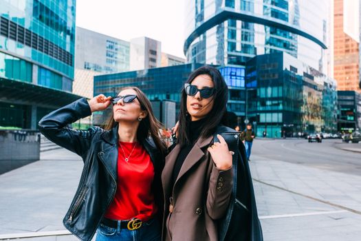Two stylish women wearing sunglasses and chic outfits pose confidently in a modern urban setting.