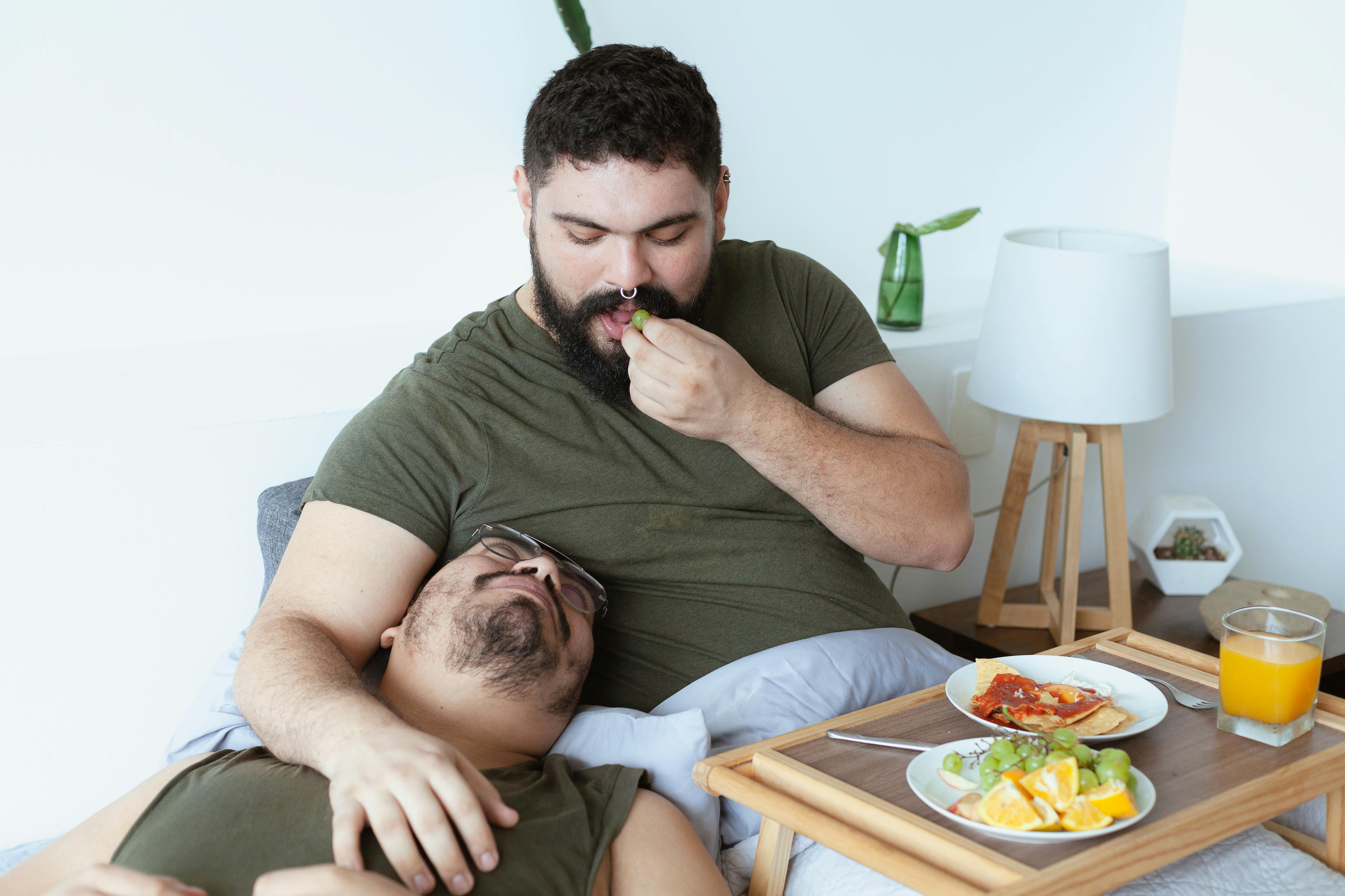 a bearded man eating grapes beside a man lying on bed