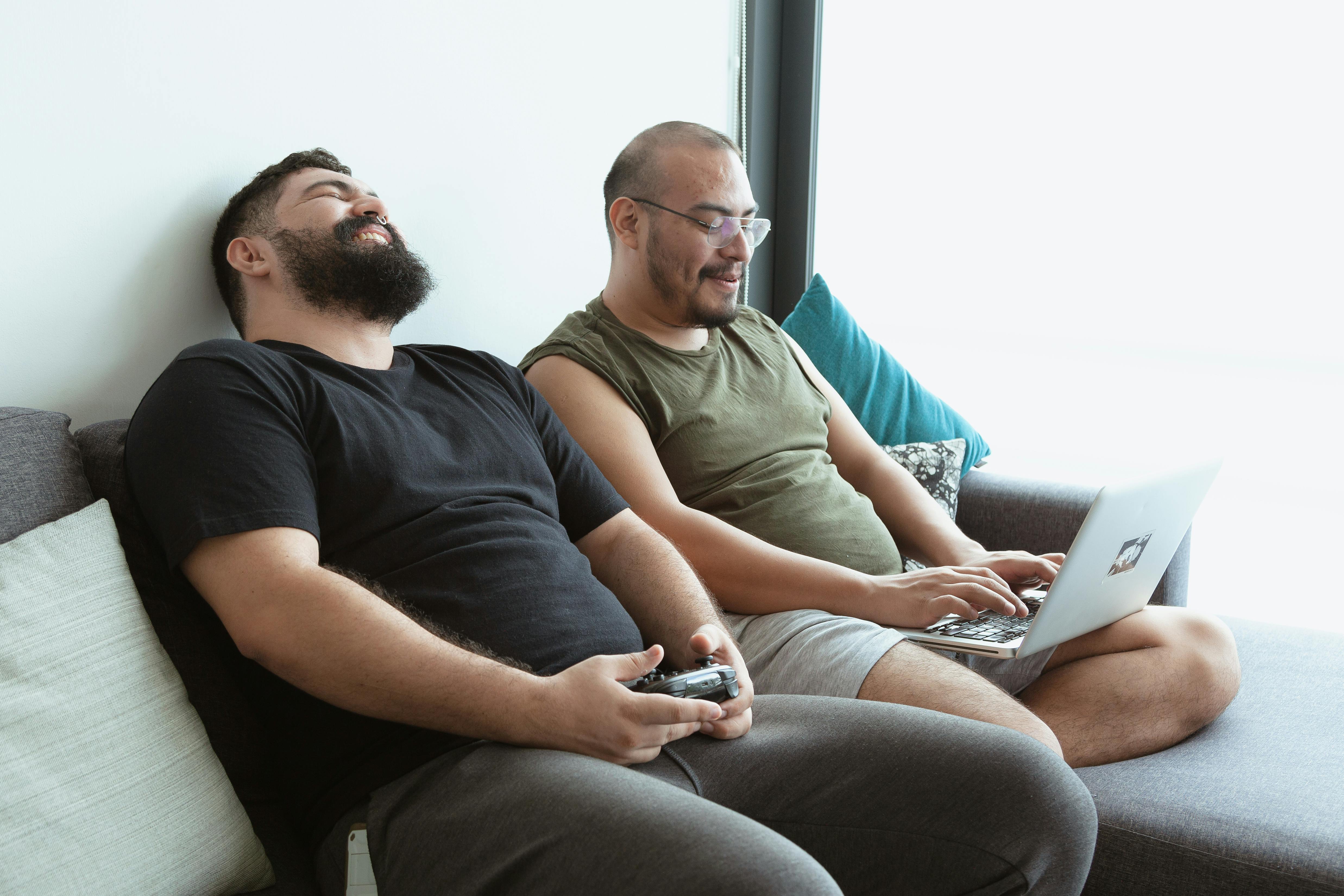 a man using a laptop sitting beside a man holding a game controller