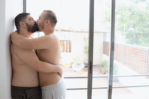 Photo of Shirtless Men Kissing Near a Glass Door