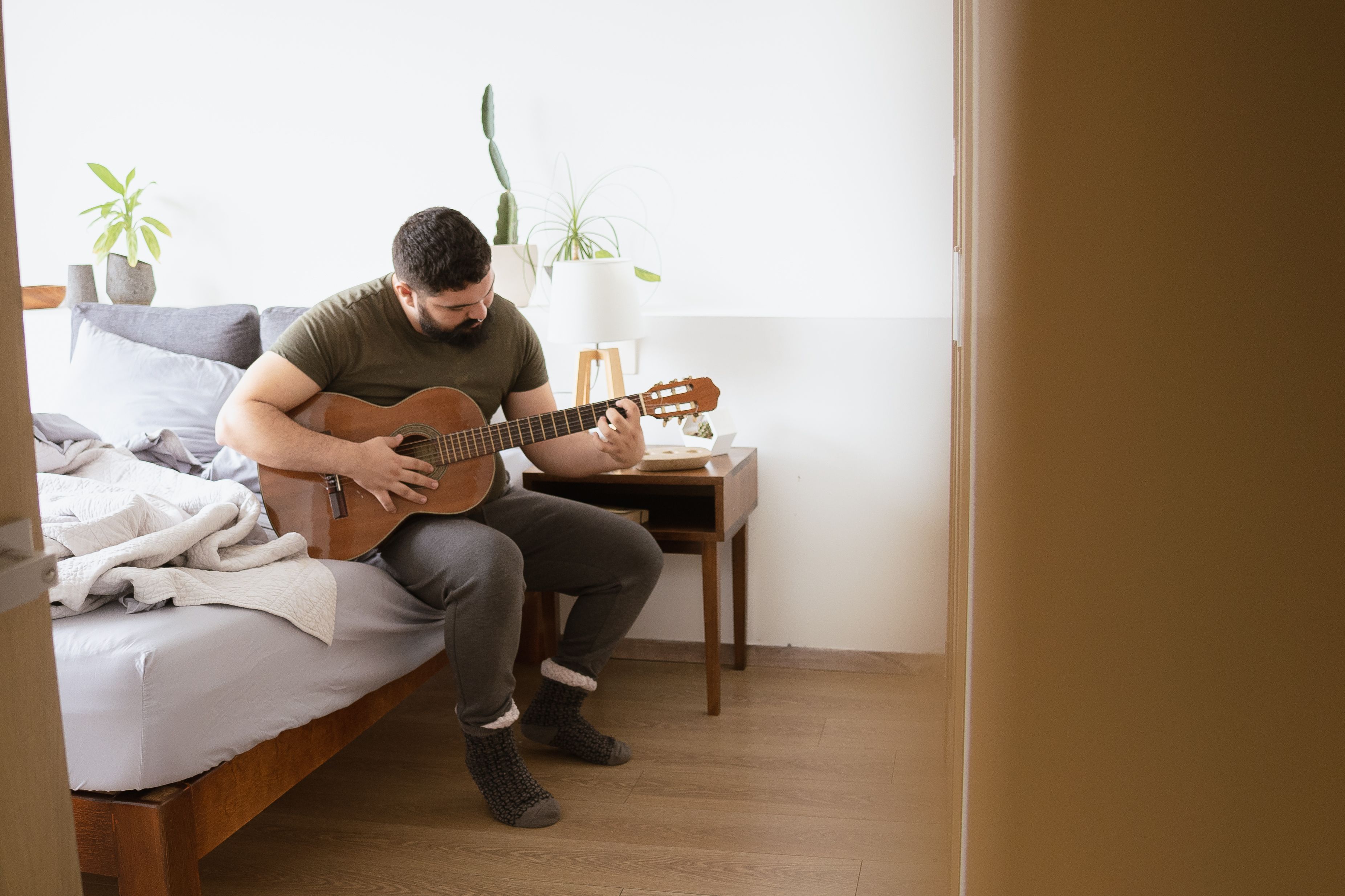 A man sitting on the edge of a bed at home, practicing chords on an acoustic guitar
