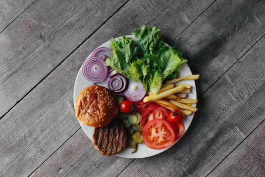 Delicious flat lay of burger components including bun, lettuce, tomatoes, onions, and fries on rustic wood.