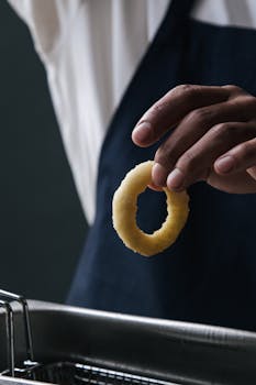 Close-up of a chef holding a raw onion ring above a deep fryer, ready to cook.