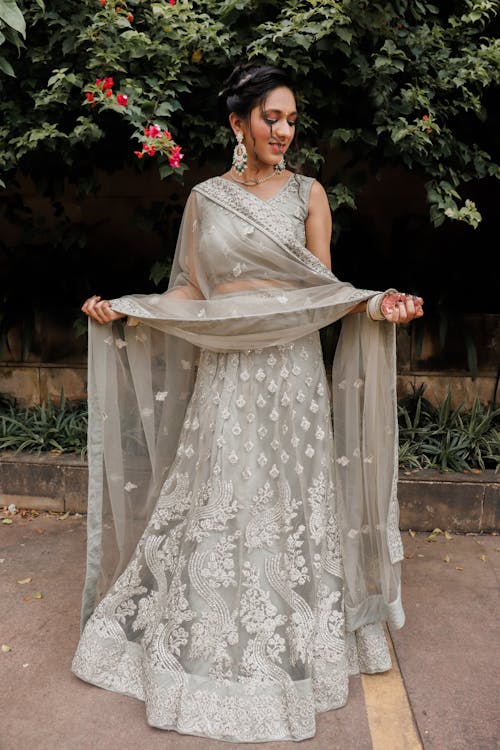 Full body of elegant young ethnic bride with dark hair in traditional Indian lehenga dress touching dupatta shawl and looking down in green park before wedding ceremony