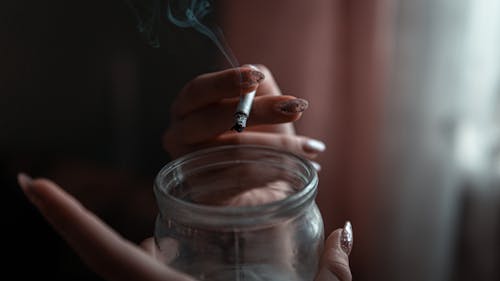 Person Holding Clear Glass Jar and a Cigarette
