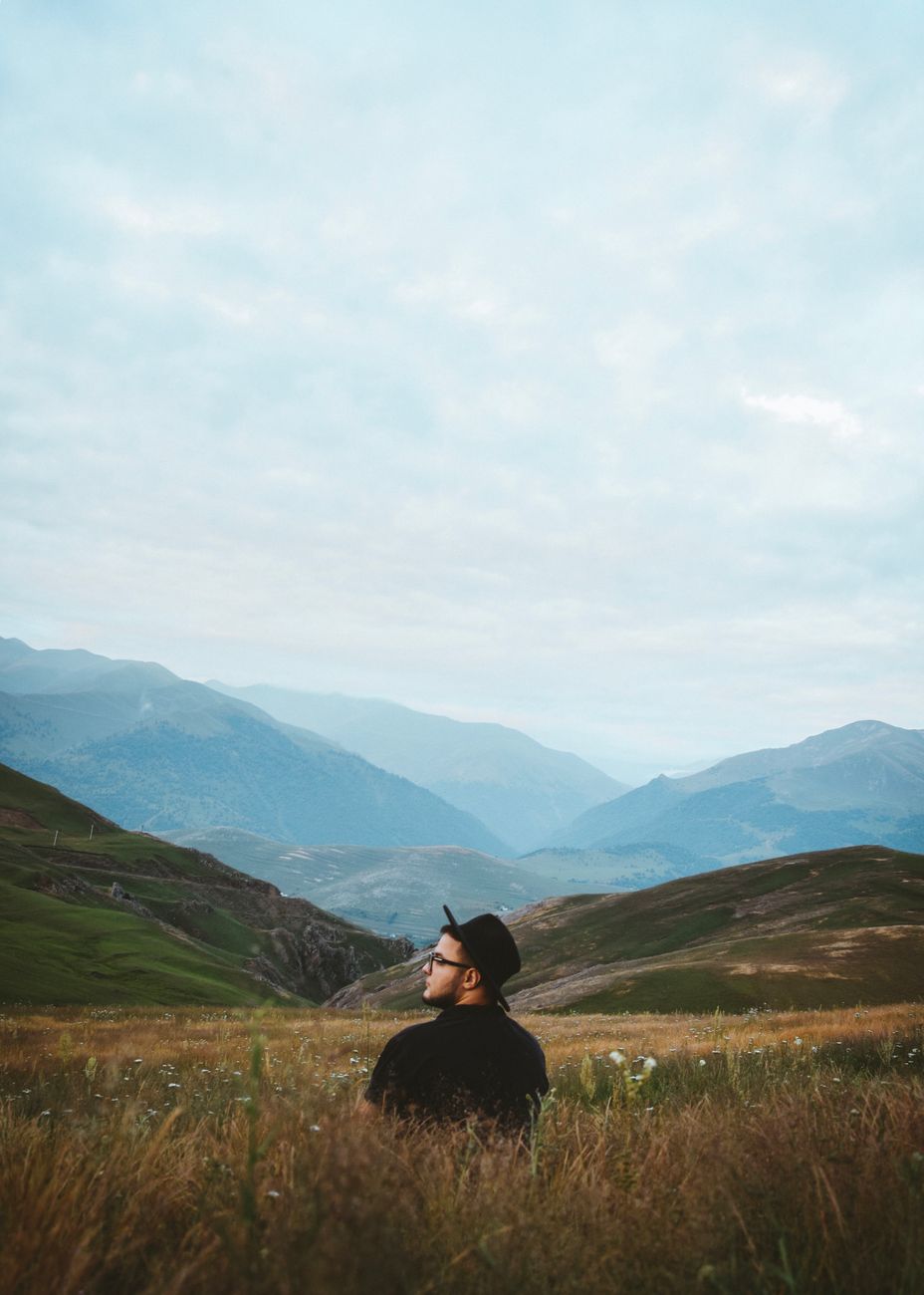 A man wearing a hat sits peacefully in a grassy field with scenic mountain views.