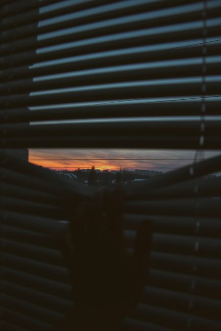 A silhouette of a hand at a window with blinds, capturing a dramatic sunset horizon.