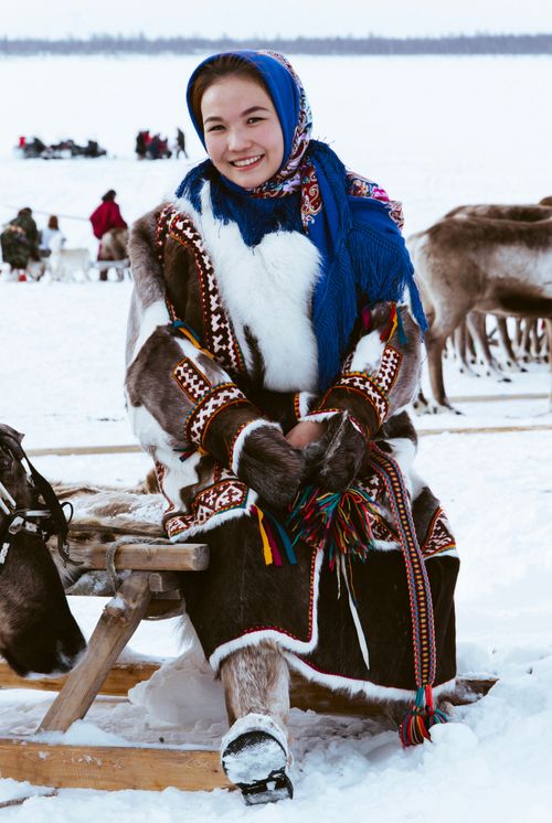 Free A smiling woman wearing traditional winter clothing sits on a sled amidst snow and reindeer. Stock Photo