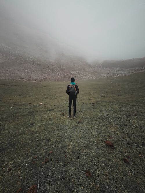 Person in Black Jacket and Black Pants Standing on Grass Field Looking at a Foggy Sky