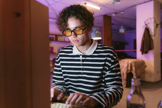 A young man wearing sunglasses types on a retro computer keyboard in a cozy indoor workspace.