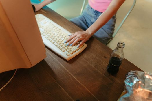 A person with blue manicured nails using a computer keyboard on a wooden table with snacks and a drink.