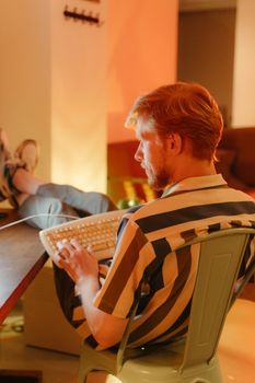 A young man with facial hair sitting indoors, typing on a vintage keyboard in relaxed setting.