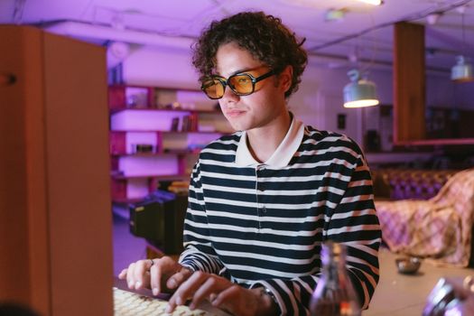 Man with curly hair in striped shirt typing on computer indoors, wearing yellow sunglasses, warm lighting.