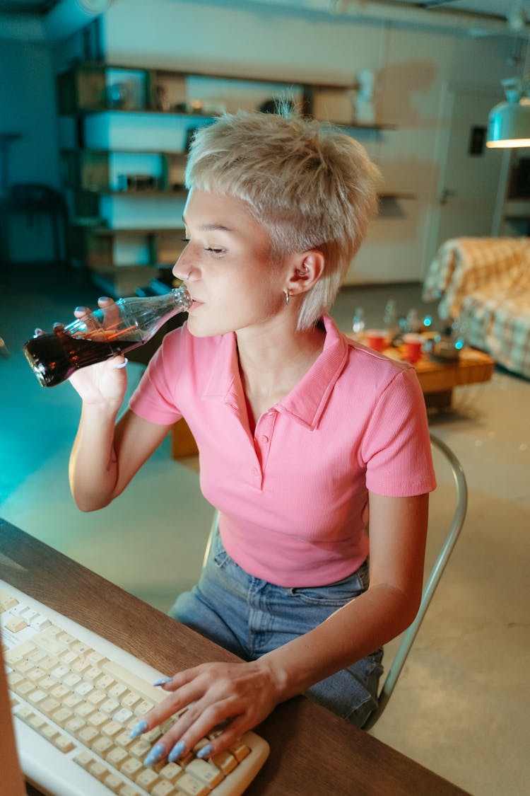 A Woman In Pink Shirt Sitting At The Work Desk While Drinking Bottled Beverage