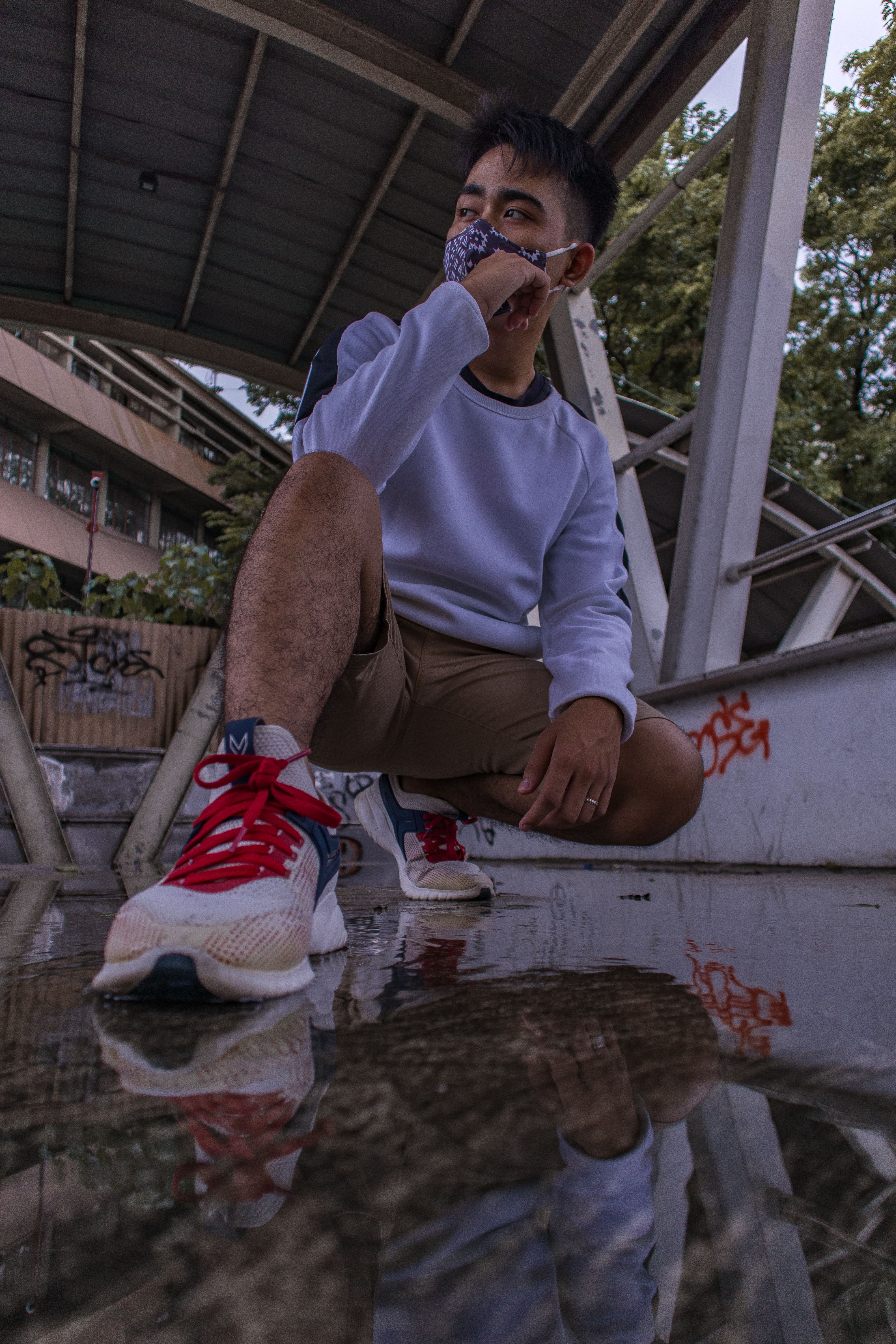 A man in casual attire poses confidently with mask in an urban setting, reflected below.