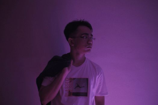 Man with eyeglasses stands in purple lighting, creating a serene, contemplative mood.