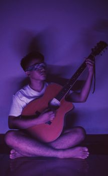 Blindfolded man sitting cross-legged playing guitar in moody purple lighting.